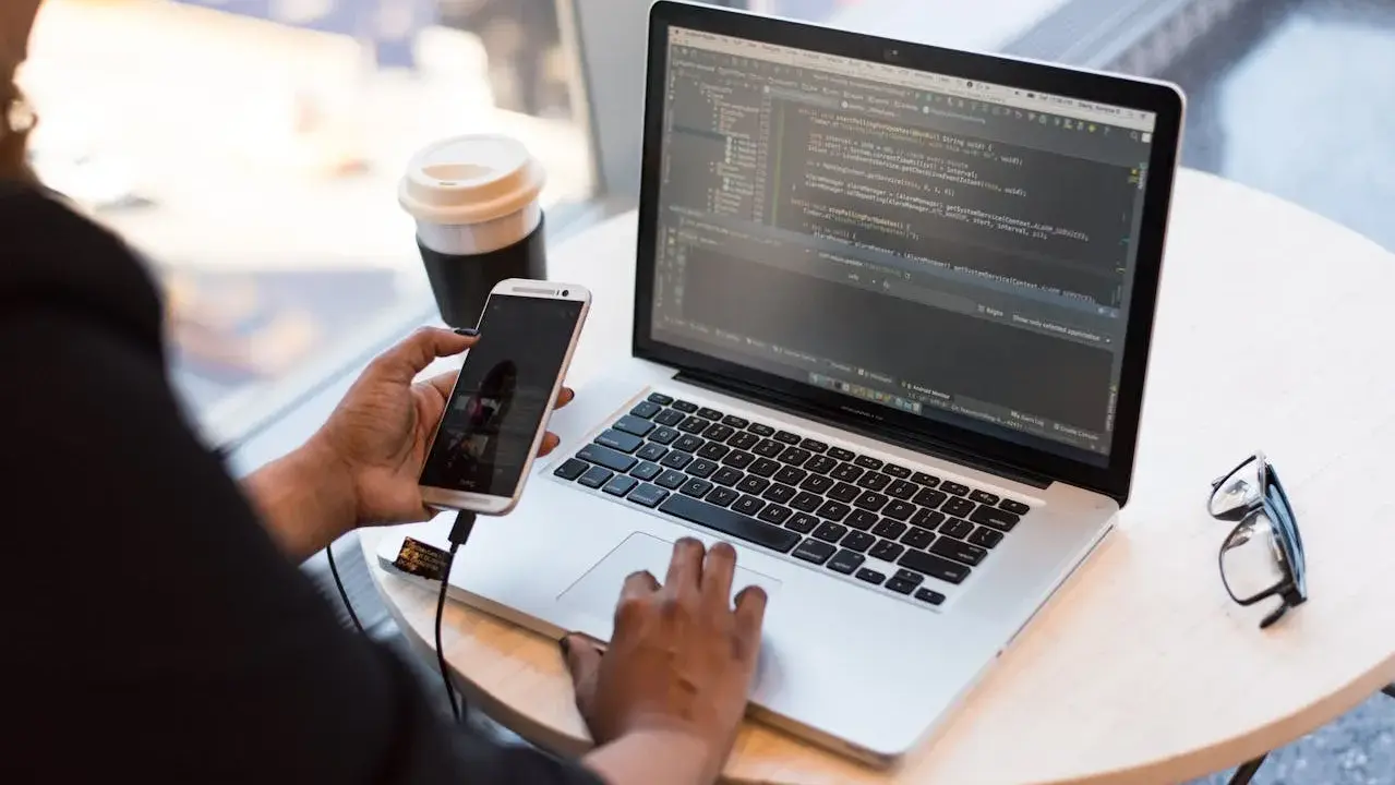 Osoba pracuje przy laptopie z kodem, używając telefonu do testowania aplikacji z **selenium**. Obok kawa i okulary.