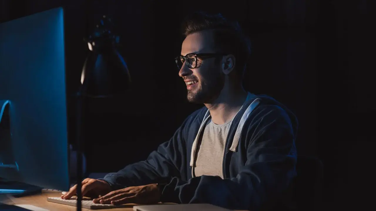 Uśmiechnięty mężczyzna w okularach pracuje nocą przy komputerze, przygotowując się do studiów podyplomowych tester oprogramowania.