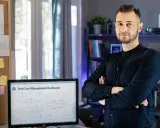 Mężczyzna w czarnej koszuli stoi obok biurka z monitorem wyświetlającym dashboard zarządzania testami. Tablica z notatkami i schematami podkreśla jego zaangażowanie w **quality engineering**.