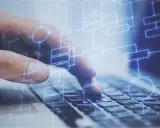 Dłoń na klawiaturze laptopa, nad nią schemat blokowy symbolizujący testy na produkcji.