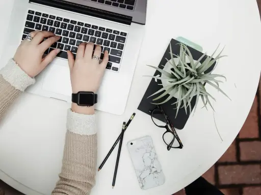Dłonie piszą na laptopie, obok leżą okulary, telefon i roślina. Może to być błąd interfejsu, gdy coś nie działa.