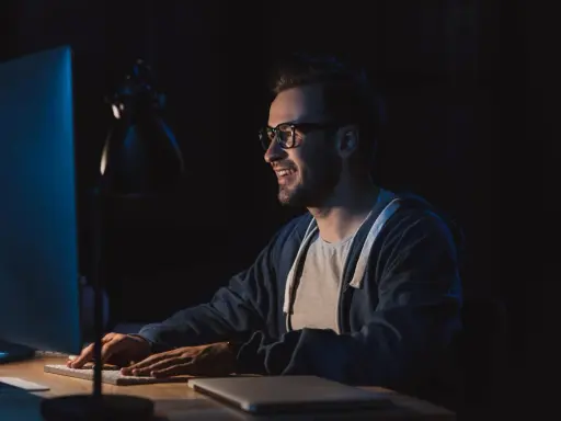 Uśmiechnięty mężczyzna w okularach pracuje nocą przy komputerze, przygotowując się do studiów podyplomowych tester oprogramowania.