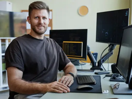 Uśmiechnięty qa engineer siedzi przy biurku z wieloma monitorami, gotowy do testowania oprogramowania.