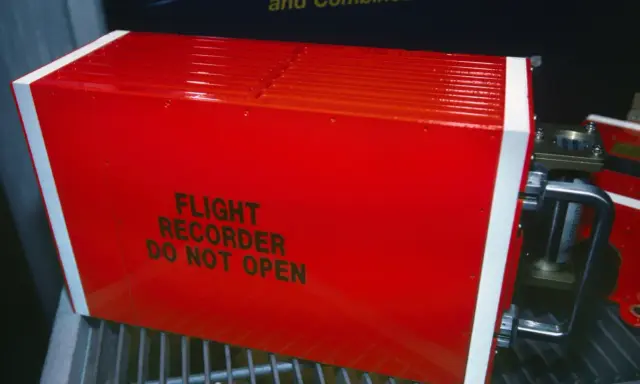 Czerwony, jaskrawy rejestrator lotu, znany jako czarna skrzynka inaczej, z napisem "FLIGHT RECORDER DO NOT OPEN".