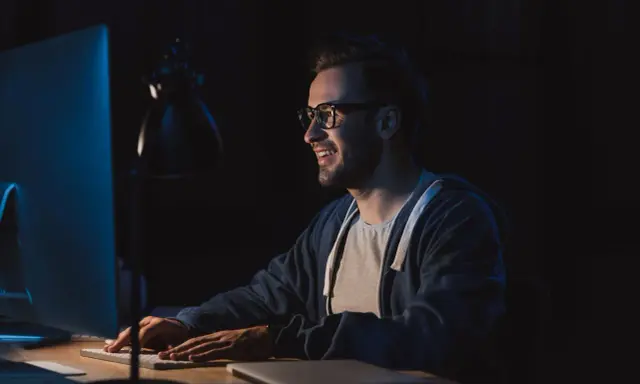 Uśmiechnięty mężczyzna w okularach pracuje nocą przy komputerze, przygotowując się do studiów podyplomowych tester oprogramowania.