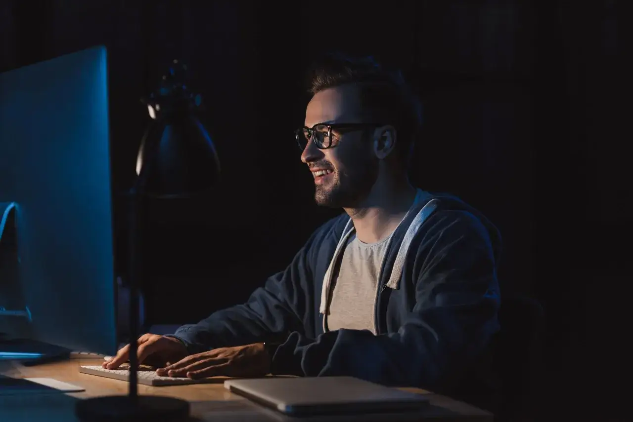 Mężczyzna w okularach z uśmiechem pracuje przy komputerze, ucząc się do studi&oacute;w podyplomowych tester oprogramowania.